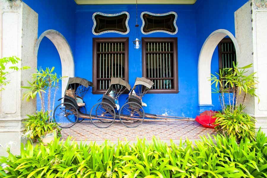 Tres rickshaws tradicionales estacionados frente a un edificio azul vibrante con ventanas arqueadas ornamentadas en un entorno tropical soleado.