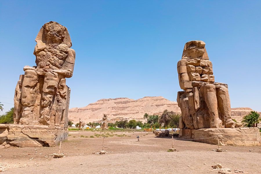 Dos enormes estatuas de piedra, los Colosos de Memnón, se alzan en un paisaje desértico con un fondo de montañas bajo un cielo azul brillante.