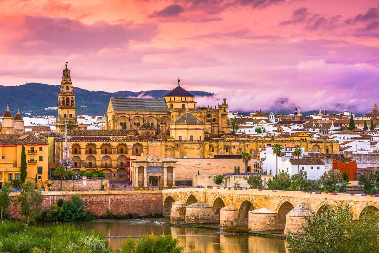 Vista de Córdoba, España al atardecer, con la Mezquita-Catedral dominando el horizonte, el puente de piedra sobre el río y el cielo colorido.