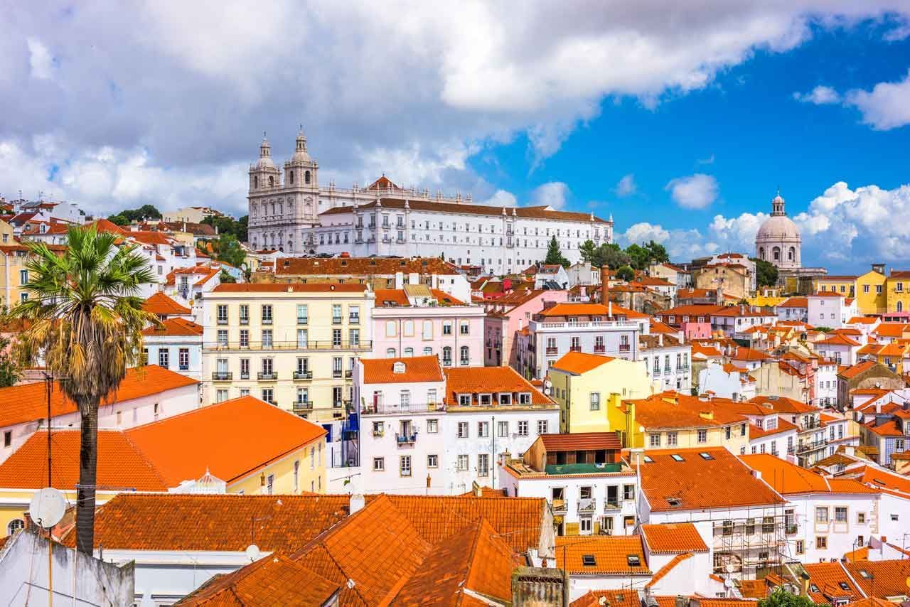 Paisaje urbano de Lisboa, Portugal, con techos de tejas naranjas, edificios blancos y una gran iglesia bajo un cielo azul brillante con nubes.