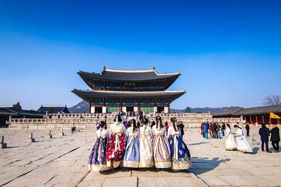 Personas con coloridos vestidos tradicionales coreanos Hanbok posan frente al Palacio Gyeongbokgung, Seúl, Corea del Sur, bajo un cielo azul claro.