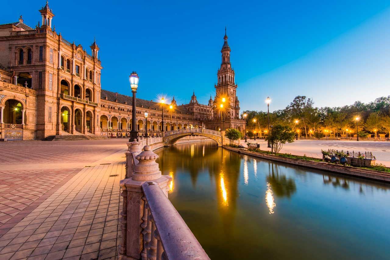 Plaza de España en Sevilla, España, al anochecer con edificios iluminados reflejados en el canal.