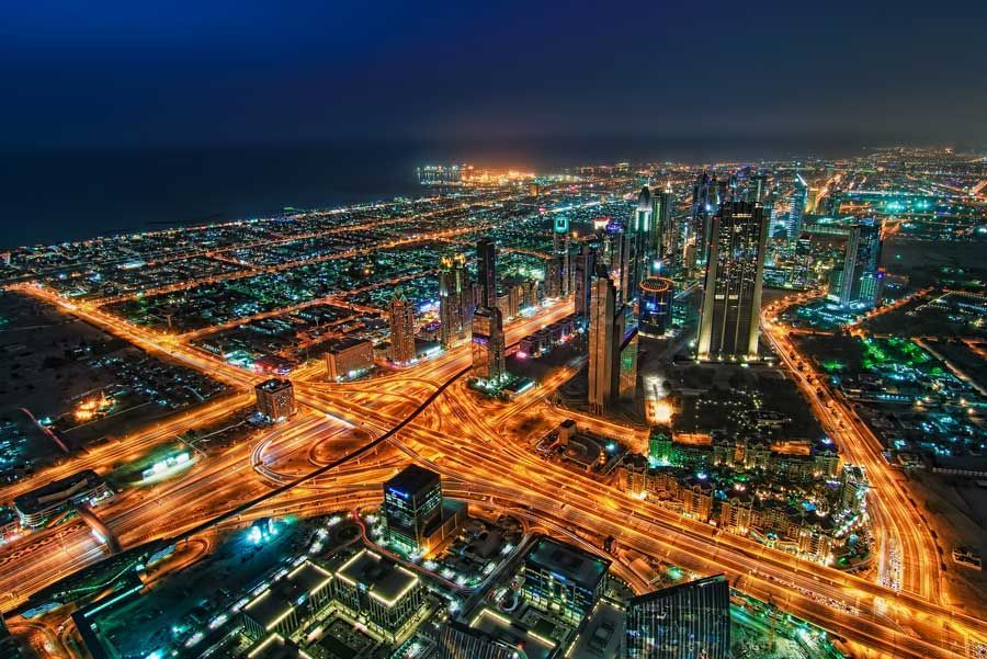 Vista aérea nocturna de Dubái. Paisaje urbano iluminado con brillantes luces naranjas y el océano azul oscuro de fondo.