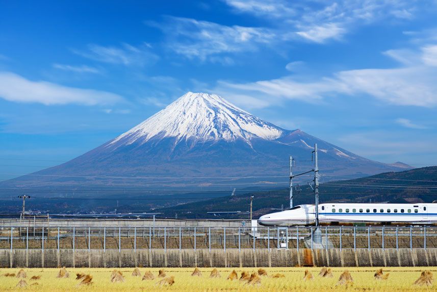 Un tren Shinkansen pasa a toda velocidad por un campo dorado con el monte Fuji al fondo bajo un cielo azul brillante.