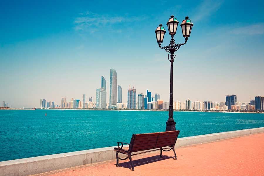 Un banco del parque y una farola ornamentada miran al mar azul y al horizonte de la ciudad en un día soleado.