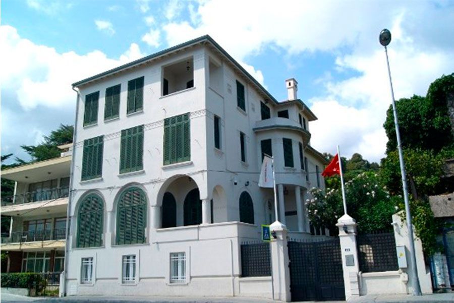 Edificio blanco con persianas verdes y bandera turca, probablemente un consulado o embajada, en un entorno exterior soleado.