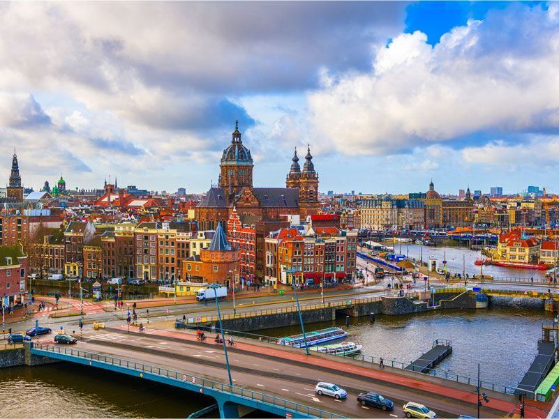 Paisaje urbano de Ámsterdam con una gran iglesia, puentes, canales y edificios coloridos bajo un cielo nublado.