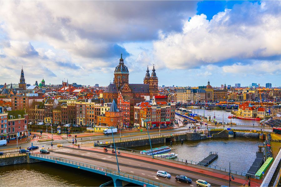 Paisaje urbano de Ámsterdam con canales, puentes y la Iglesia de San Nicolás bajo un cielo nublado. Se ven coloridos edificios y barcos.