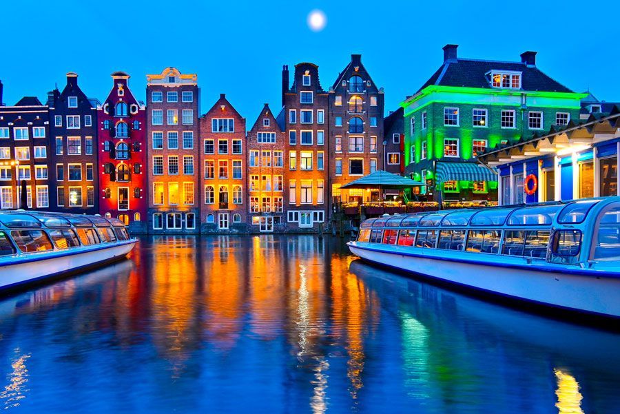Canal de Ámsterdam al anochecer, edificios coloridos iluminados a lo largo del agua, barcos turísticos atracados, luna en el cielo.