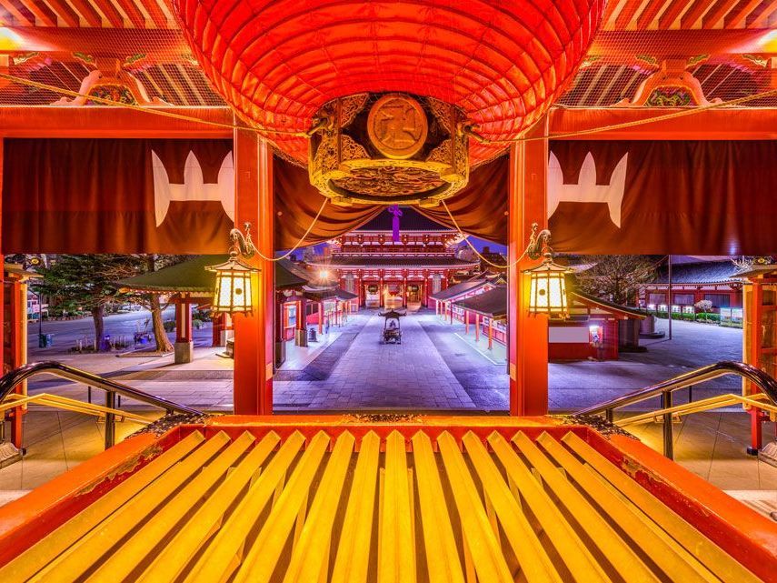 Un vibrante templo japonés rojo y dorado al anochecer, visible a través de una puerta. El templo está iluminado con faroles.