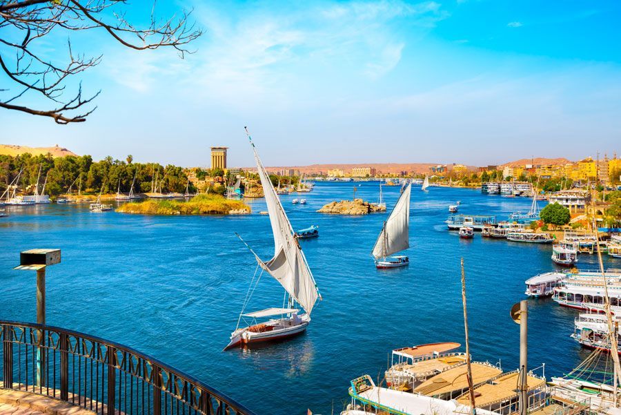 Barcos con velas blancas en el azul río Nilo en Asuán, Egipto, en un día soleado. Edificios y árboles bordean las orillas.