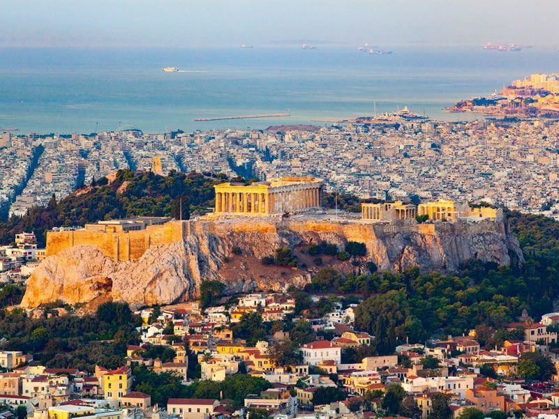 La Acrópolis de Atenas, Grecia, iluminada por el sol, se encuentra en la cima de una colina rocosa con vistas a una ciudad densamente construida y al mar.