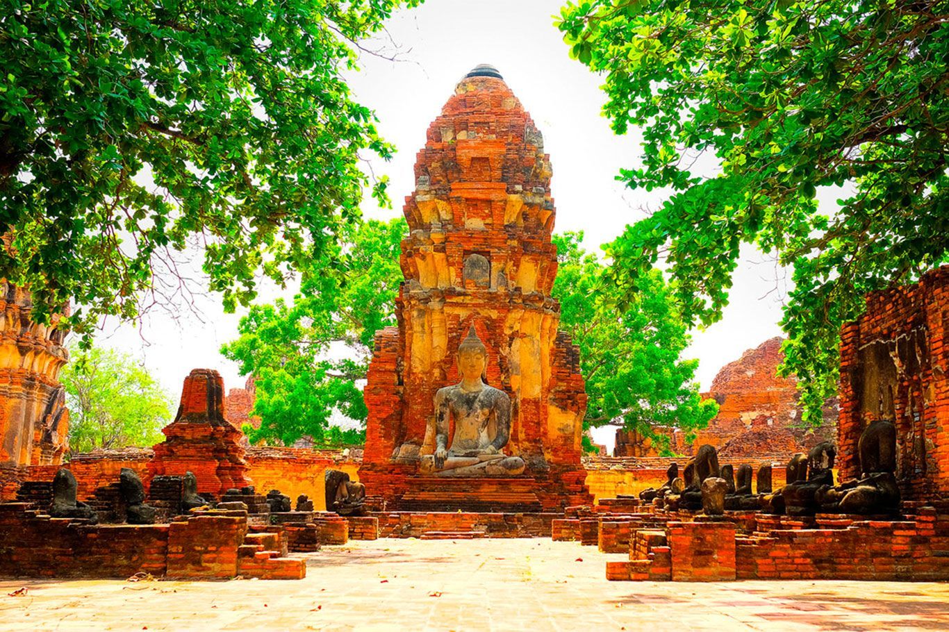 Ruinas de un antiguo templo en Ayutthaya, Tailandia. Una estructura de ladrillo erosionado con una estatua de Buda sedente está rodeada de exuberante vegetación.