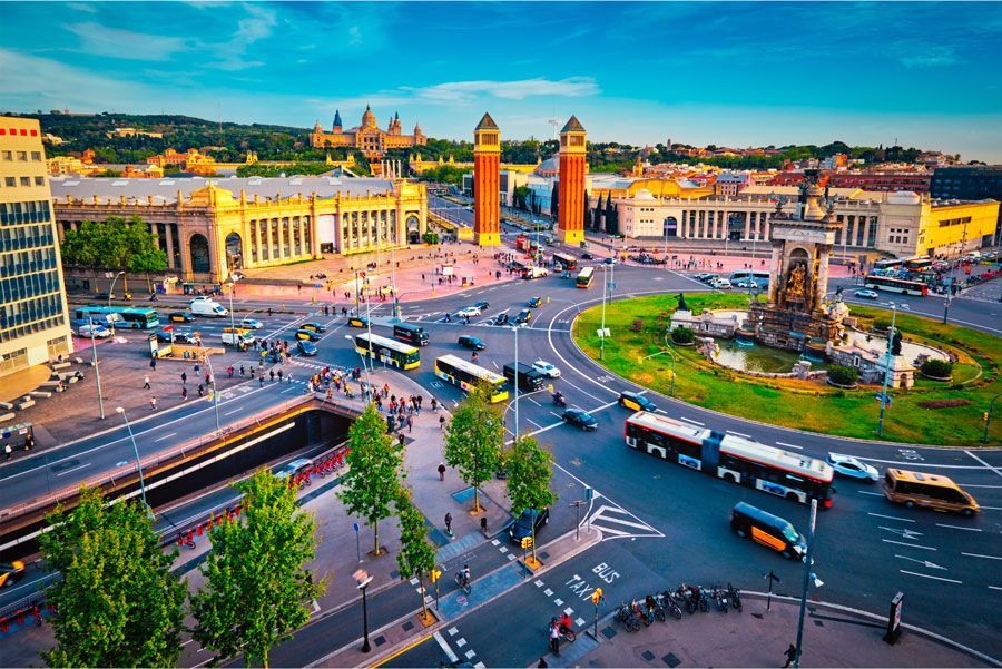 Vista aérea de la Plaza de España, Barcelona. Rotonda concurrida con vehículos y peatones, flanqueada por edificios monumentales de tonos cálidos.