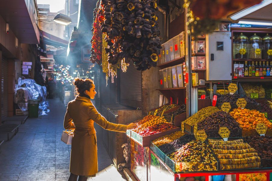 Una mujer con abrigo color canela mira productos secos en un colorido puesto de mercado. La luz del sol se filtra por un estrecho callejón en Turquía.