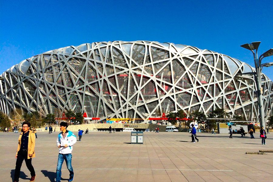 Estadio Nido de Pájaro en Pekín, China, con una compleja fachada de estructura de acero bajo un cielo azul claro; la gente camina al frente.