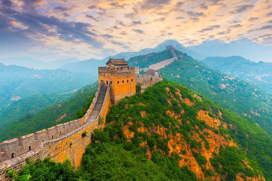 La Gran Muralla China se extiende por la cresta de una montaña bajo un cielo colorido. Una torre de vigilancia se alza en la cima, con secciones de la muralla visibles.