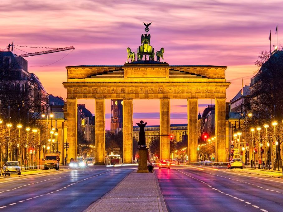Puerta de Brandeburgo iluminada al anochecer, Berlín. Estructura dorada con una cuadriga en su cima, flanqueada por farolas y tráfico en una amplia avenida.