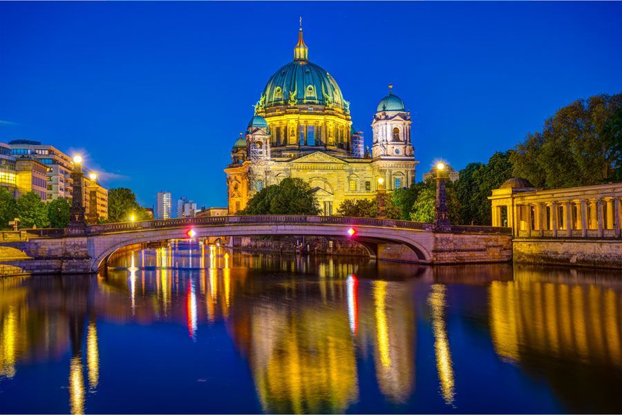La Catedral de Berlín de noche, reflejada en el río Spree. Cúpula verde azulado, luces amarillas en el puente y edificios bajo un cielo azul oscuro.