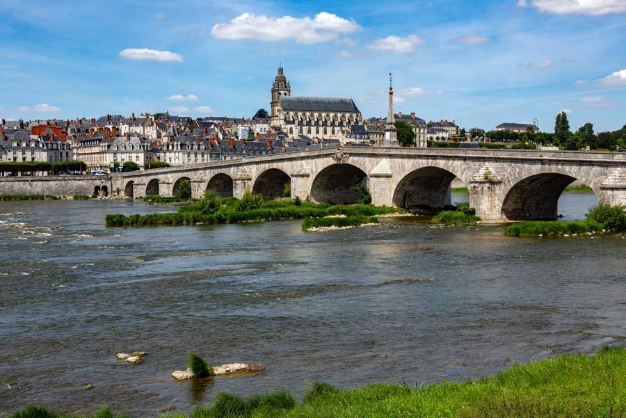Puente de arco sobre un río ancho, con el horizonte de la ciudad y una iglesia al fondo bajo un cielo azul.