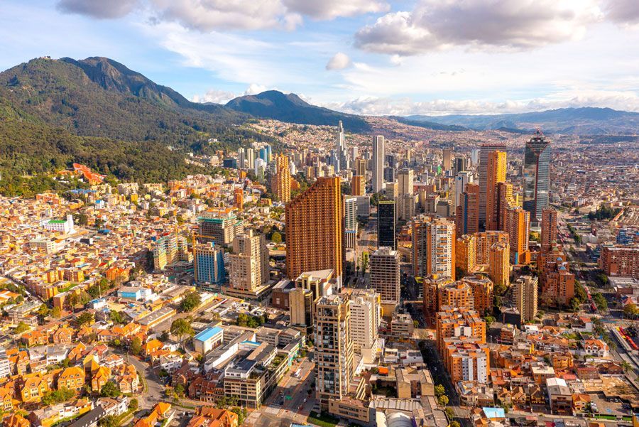 Vista aérea de Bogotá, Colombia, con un denso paisaje urbano con edificios de gran altura, barrios circundantes y montañas al fondo bajo un cielo azul.