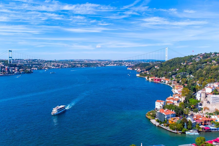 Vista aérea del estrecho del Bósforo en Estambul, Turquía. Un barco blanco navega por las aguas azules con un puente y una ciudad a ambos lados bajo un cielo despejado.