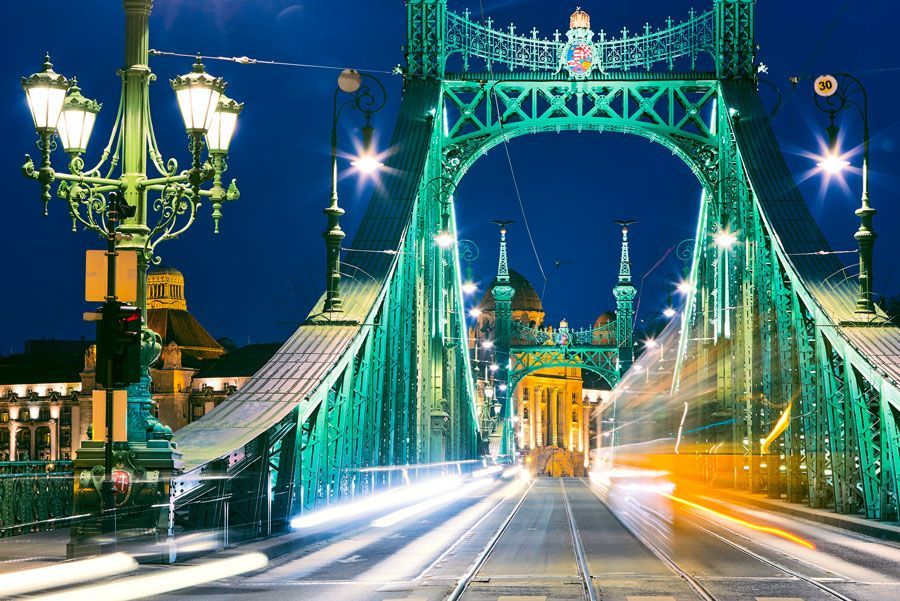 El Puente de la Libertad en Budapest, iluminado de verde por la noche, con luces de automóviles y edificios borrosos al fondo.