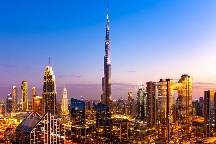 El horizonte de Dubái al atardecer, con el Burj Khalifa como rascacielos central iluminado. Otros edificios modernos lucen tonos dorados y naranjas.