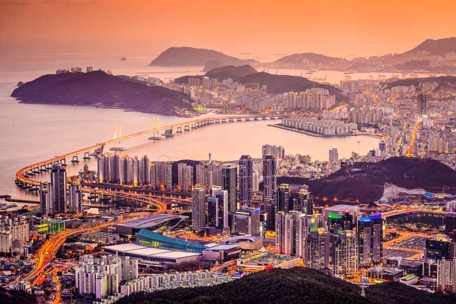 Paisaje urbano de Busan, Corea del Sur, al atardecer con un puente sobre el agua, montañas y edificios iluminados.