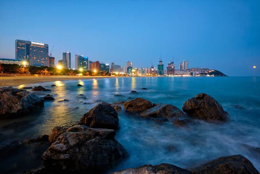 Horizonte de la ciudad frente a la playa al anochecer, con rocas en primer plano y edificios con luces reflejándose en el agua.