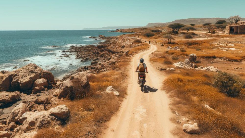 Persona conduciendo una bicicleta por un camino de tierra a lo largo de una costa rocosa, océano a la izquierda, paisaje árido.