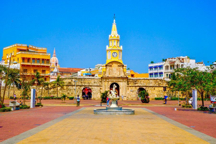 Paisaje urbano de Cartagena, Colombia, con la puerta de la Torre del Reloj, edificios amarillos vibrantes, cielo azul y una fuente.