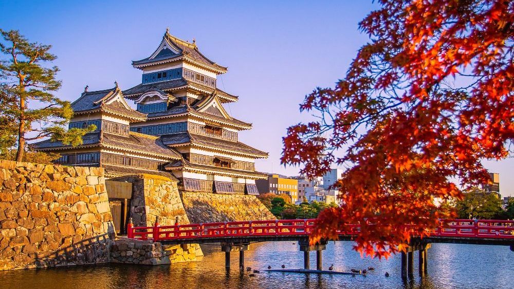 Castillo de Matsumoto en Japón con un puente rojo, rodeado de agua y follaje otoñal.