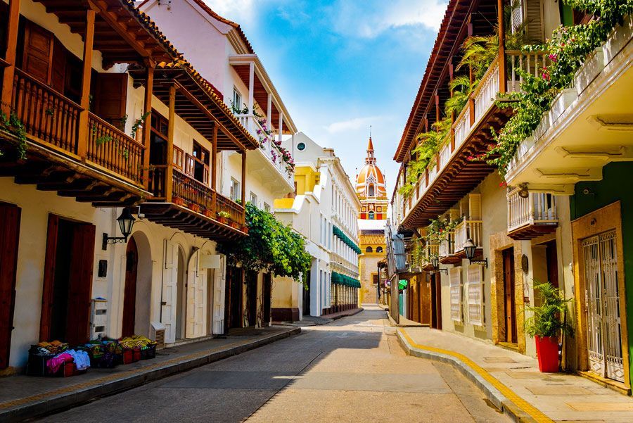 Calle adoquinada en Cartagena, Colombia, bordeada de coloridos edificios coloniales con balcones y la cúpula de una iglesia visible en la distancia.