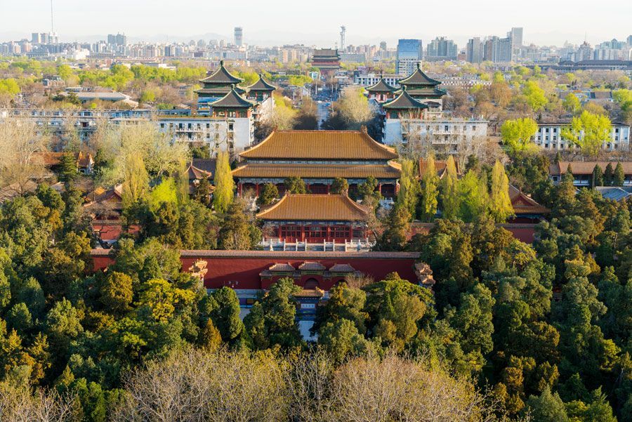 Vista aérea de la Ciudad Prohibida en Pekín, China, con edificios de tejados dorados rodeados de frondosos árboles. Al fondo, se ve el horizonte de la ciudad.