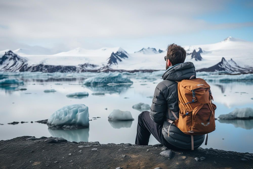 Un hombre con una mochila naranja se sienta junto a un lago glacial, mirando icebergs y montañas cubiertas de nieve.
