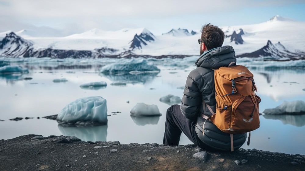 Hombre con mochila naranja sentado y mirando un lago glaciar con icebergs y montañas nevadas.