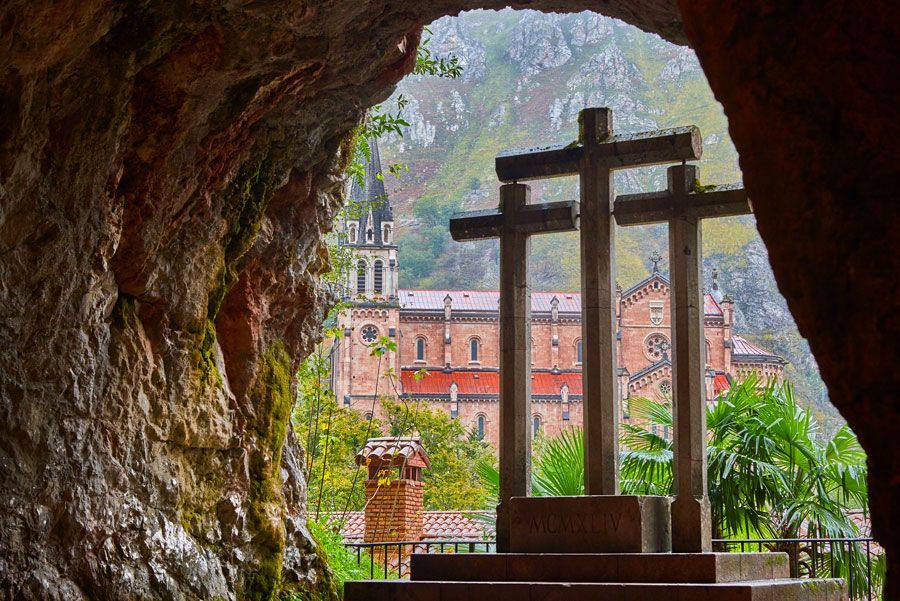 Tres cruces frente a una catedral, vistas desde la entrada de una cueva. Un paisaje verde rodea las estructuras; colores apagados.
