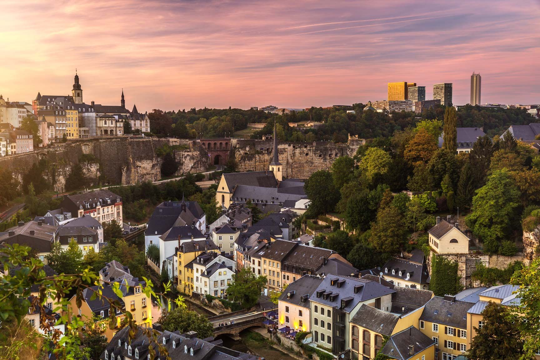El horizonte de la ciudad de Luxemburgo al atardecer, con edificios ubicados a lo largo de un valle y un cielo rosa y naranja.