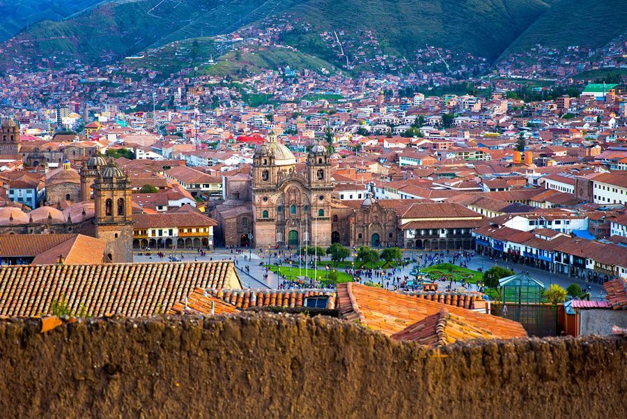 Paisaje urbano de Cusco, Perú, con una gran catedral en la plaza central, rodeada de techos de tejas rojas y un fondo montañoso.