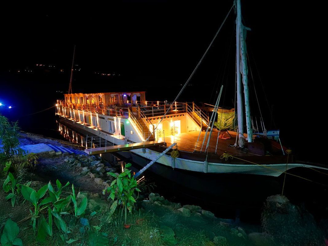 Barco iluminado de noche, atracado cerca de una orilla con vegetación. Se ven edificios desde el barco.
