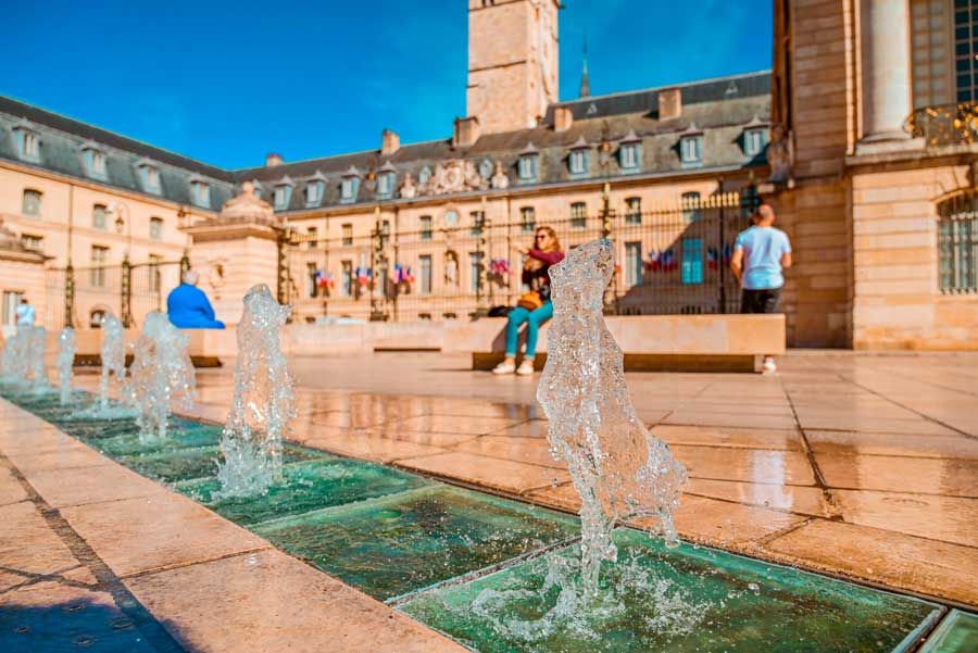 Las fuentes rocían agua en una plaza frente a un edificio europeo. La gente se relaja en los bancos cercanos.