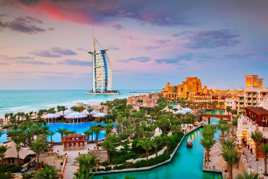 El horizonte de Dubái muestra el hotel Burj Al Arab y un complejo con piscinas y canales, bajo un cielo colorido al atardecer.