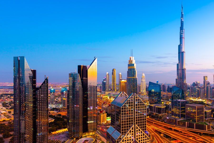 Paisaje urbano de Dubai al anochecer con numerosos rascacielos, incluido el Burj Khalifa, iluminados contra un cielo azul y naranja.