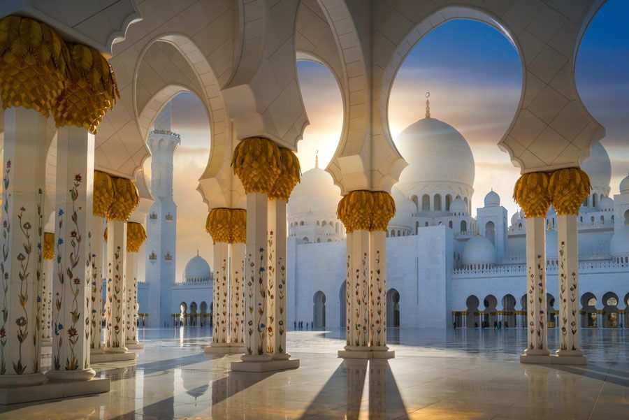 Vista interior de la Gran Mezquita Sheikh Zayed en Abu Dhabi, con columnas blancas, acentos dorados y las cúpulas del edificio bajo un cielo al atardecer.