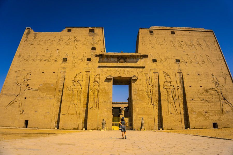 Antiguo templo egipcio de Horus en Edfu, con una persona caminando hacia la entrada bajo un cielo azul despejado. Las paredes son de color canela con jeroglíficos tallados.