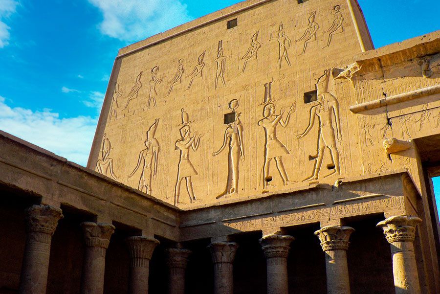 Vista exterior del Templo de Edfu en Egipto, con muros de arenisca con jeroglíficos tallados. La luz del sol ilumina la estructura contra un cielo azul.