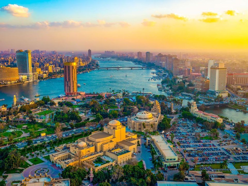 Vista aérea de El Cairo, Egipto, con el río Nilo atravesando la ciudad, edificios y vegetación bajo una cálida puesta de sol de color amarillo anaranjado.