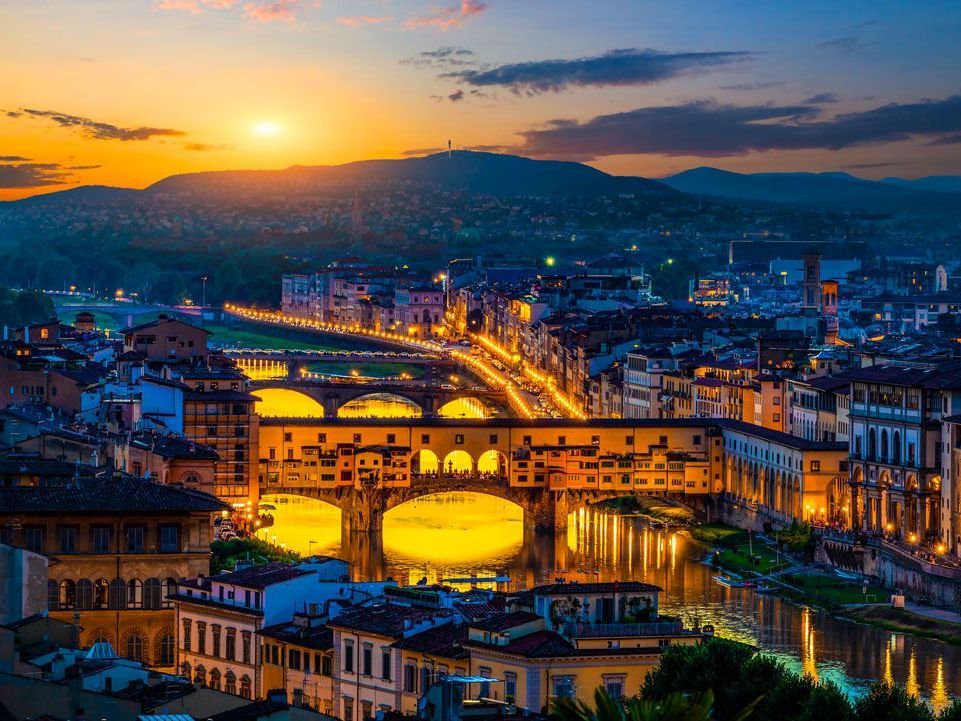 Puesta de sol sobre Florencia, Italia, con el puente Ponte Vecchio iluminado y los edificios a lo largo del río Arno reflejando las luces.