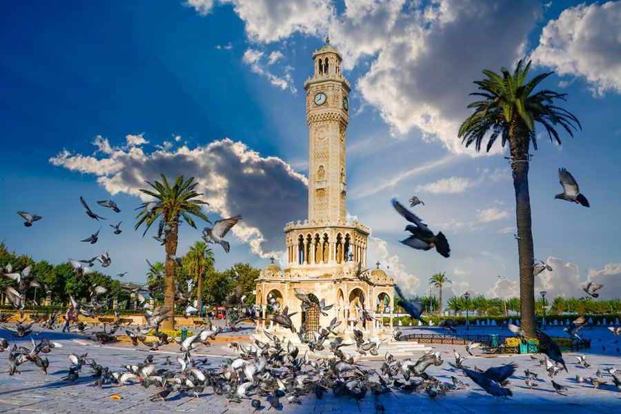 Torre del Reloj de Izmir en Turquía, rodeada de palomas y palmeras bajo un cielo azul con nubes.